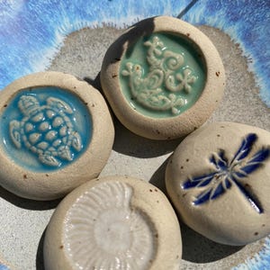 May include: Four ceramic stones with embossed designs: a turtle, a dragonfly, a sea creature, and a spiral fossil. The stones are set on a blue and white ceramic plate. The designs are in shades of blue and green.