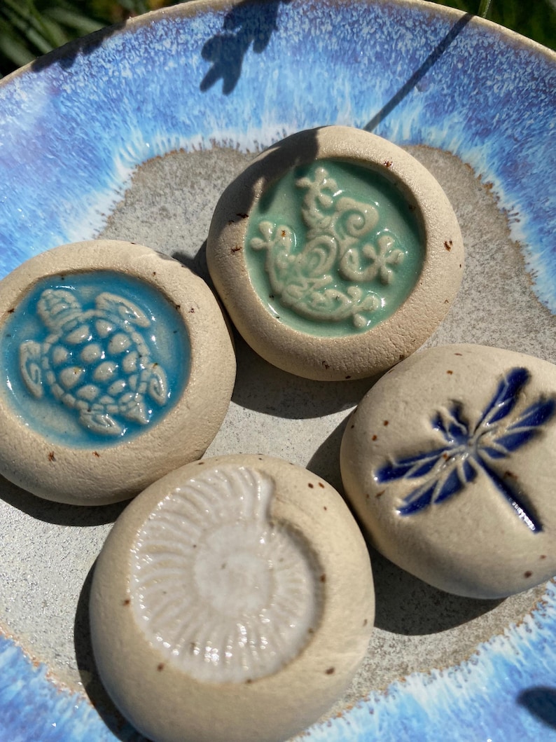 May include: Four ceramic stones with embossed designs: a turtle, a dragonfly, a sea creature, and a spiral fossil. The stones are set on a blue and white ceramic plate. The designs are in shades of blue and green.