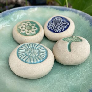 May include: Four round, off-white ceramic stones with embossed, glazed designs resting in a light green and blue crackle-glazed bowl. The designs include a white floral pattern, a blue jellyfish, a light blue sunburst, and a green ginkgo leaf.