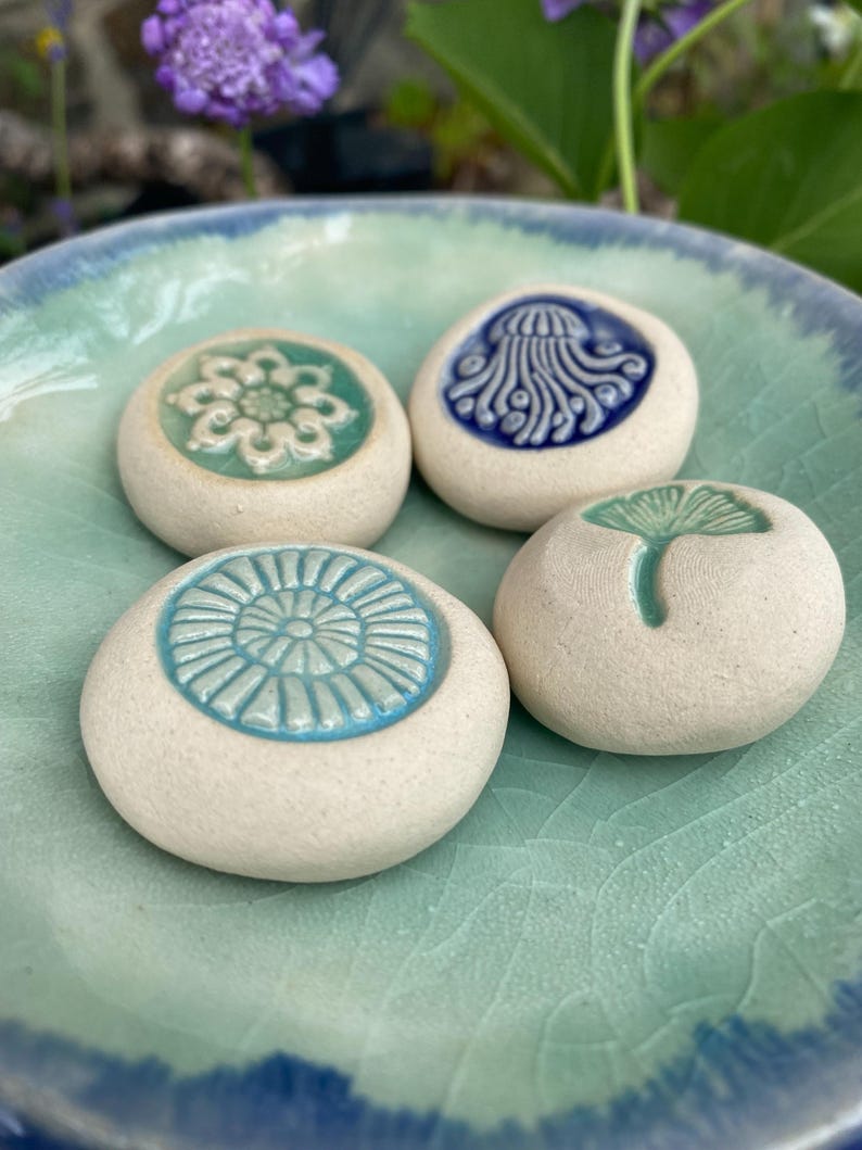 May include: Four round, off-white ceramic stones with embossed, glazed designs resting in a light green and blue crackle-glazed bowl. The designs include a white floral pattern, a blue jellyfish, a light blue sunburst, and a green ginkgo leaf.
