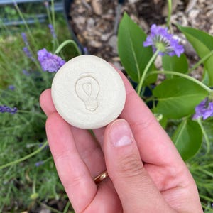 May include: A round, off-white ceramic stone featuring a debossed lightbulb icon with a heart-shaped filament in the center. The surface of the stone is smooth and matte, with a circular shape and a slightly raised edge around the central design.