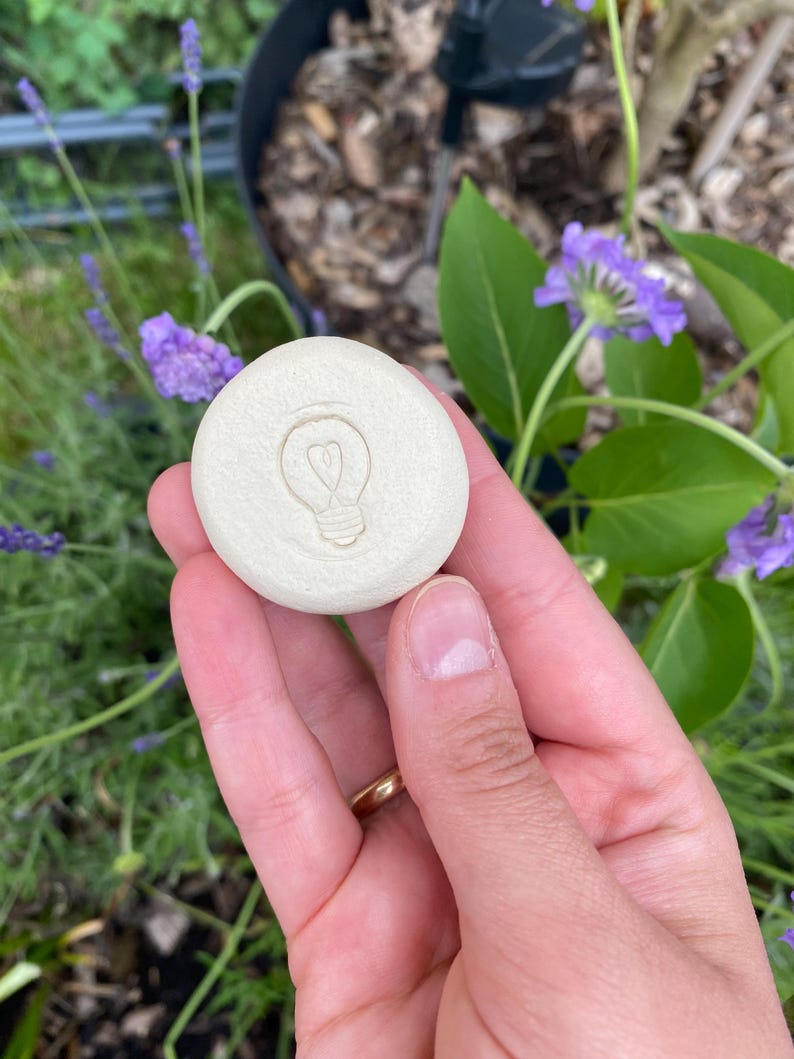 May include: A round, off-white ceramic stone featuring a debossed lightbulb icon with a heart-shaped filament in the center. The surface of the stone is smooth and matte, with a circular shape and a slightly raised edge around the central design.