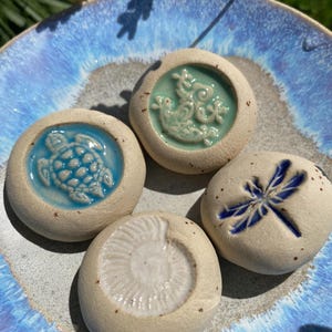 May include: Four ceramic stones with embossed designs of a turtle, a dragonfly, a seashell, and a sea creature. The stones are beige with blue and teal accents, resting on a blue and gray ceramic plate.