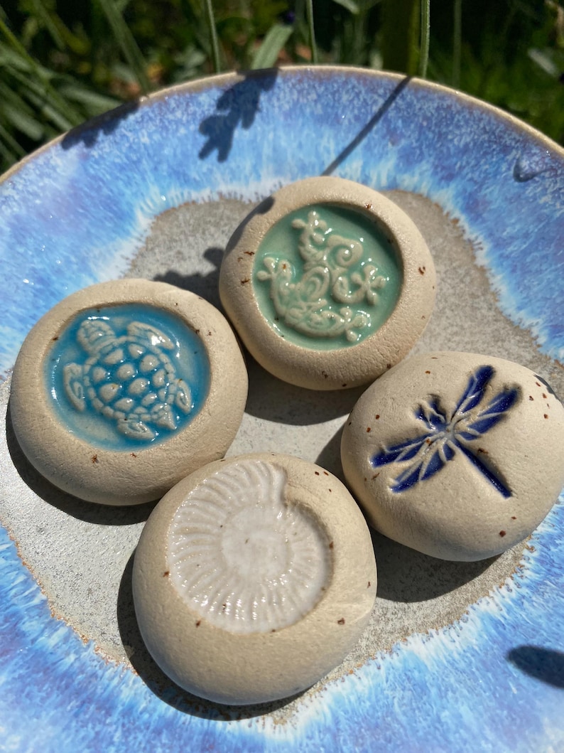 May include: Four ceramic stones with embossed designs of a turtle, a dragonfly, a seashell, and a sea creature. The stones are beige with blue and teal accents, resting on a blue and gray ceramic plate.