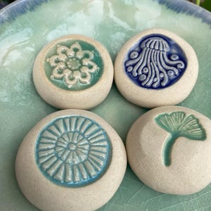 May include: Four round, off-white ceramic stones featuring embossed designs in the center. The designs include a light green flower, a dark blue jellyfish, a light blue sunburst pattern, and a light green ginkgo leaf, all resting on a bowl.