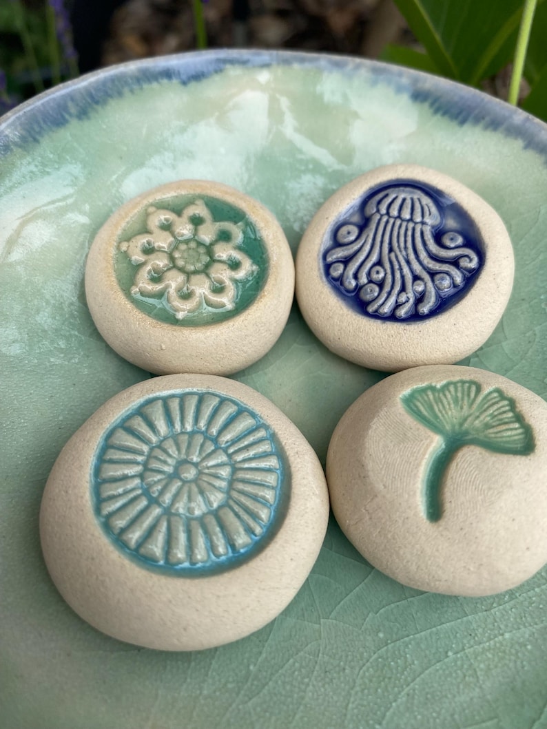 May include: Four round, off-white ceramic stones featuring embossed designs in the center. The designs include a light green flower, a dark blue jellyfish, a light blue sunburst pattern, and a light green ginkgo leaf, all resting on a bowl.