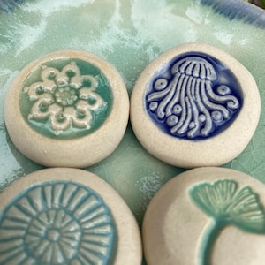 May include: Four round, off-white ceramic stones featuring embossed designs. One displays a green floral pattern, another a blue jellyfish, the third a blue radial sunburst, and the fourth a green ginkgo leaf. Each piece has a smooth, glazed finish.