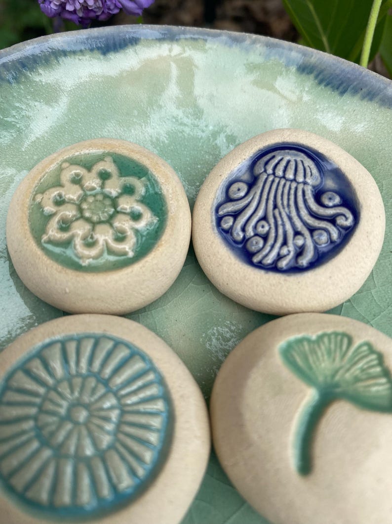 May include: Four round, off-white ceramic stones featuring embossed designs. One displays a green floral pattern, another a blue jellyfish, the third a blue radial sunburst, and the fourth a green ginkgo leaf. Each piece has a smooth, glazed finish.