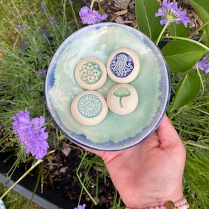 May include: A round ceramic dish with a light green and blue glazed interior containing four small, circular, off-white clay tokens. Each token features a distinct embossed design: a floral pattern, a jellyfish, a sunburst, and a ginkgo leaf.