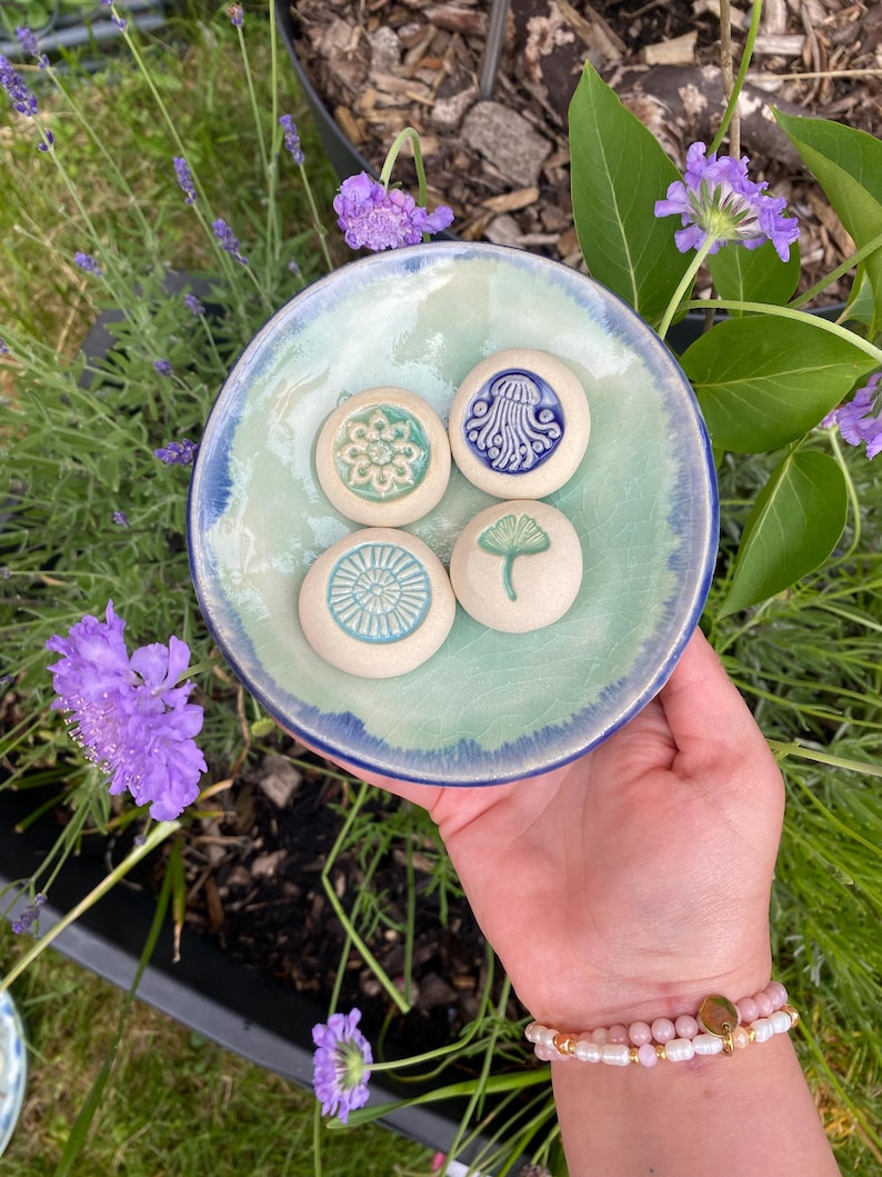 May include: A round ceramic dish with a light green and blue glazed interior containing four small, circular, off-white clay tokens. Each token features a distinct embossed design: a floral pattern, a jellyfish, a sunburst, and a ginkgo leaf.