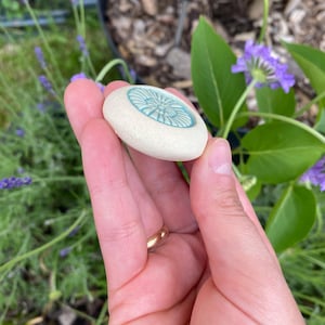 May include: A small, round, off-white ceramic stone with a textured, matte surface. The center features a circular, recessed inlay in a bright, glossy turquoise blue color, decorated with a radial, flower-like pattern of embossed lines.