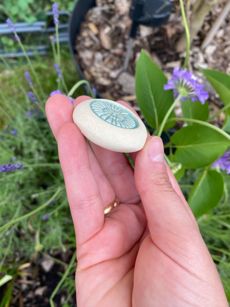 May include: A small, round, off-white ceramic stone with a textured, matte surface. The center features a circular, recessed inlay in a bright, glossy turquoise blue color, decorated with a radial, flower-like pattern of embossed lines.