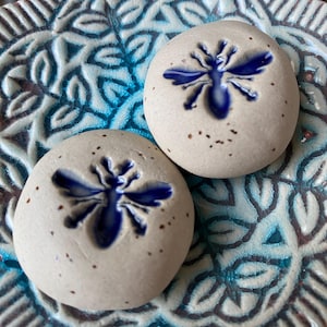 Könnte beinhalten: Zwei kleine, runde, gesprenkelte, cremefarbene Keramiksteine mit einem blauen Bienenmuster. Die Steine liegen auf einem dekorativen blau-weißen Keramikteller mit floralem Muster. Das Bienenmuster ist tiefblau.