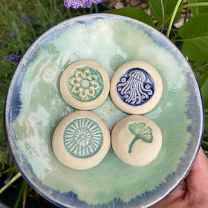 May include: A round, light green and blue glazed ceramic dish with a crackle finish, holding four small, circular, off-white ceramic tokens. Each token features a unique embossed design: a floral pattern, a jellyfish, a sunburst, and a ginkgo leaf.