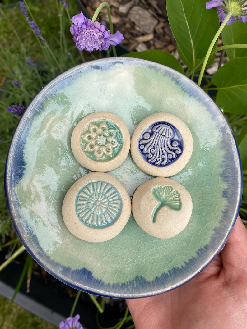 May include: A round, light green and blue glazed ceramic dish with a crackle finish, holding four small, circular, off-white ceramic tokens. Each token features a unique embossed design: a floral pattern, a jellyfish, a sunburst, and a ginkgo leaf.