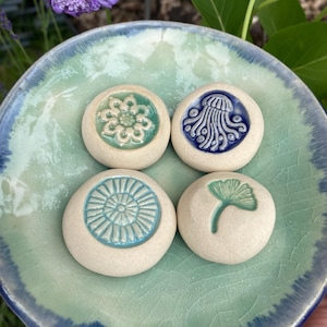 May include: Four round, off-white ceramic stones with embossed designs resting on a light green and blue glazed ceramic dish. The designs include a flower, a jellyfish, a sunburst pattern, and a ginkgo leaf, each featuring colored glaze.