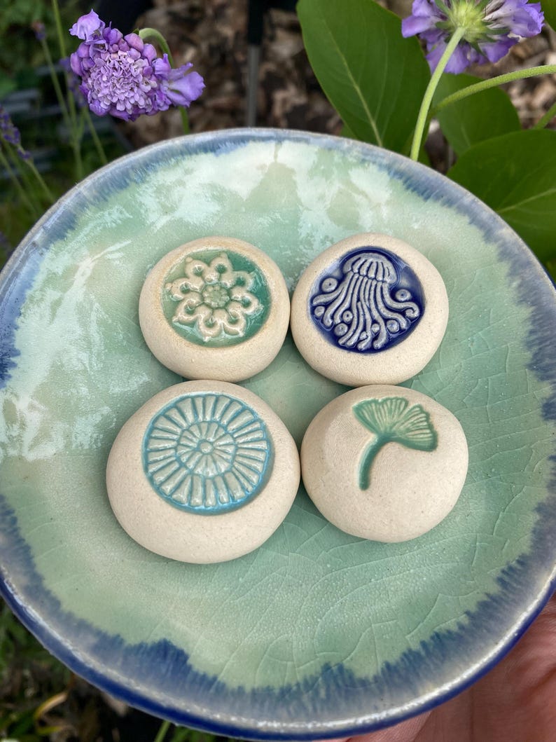 May include: Four round, off-white ceramic stones with embossed designs resting on a light green and blue glazed ceramic dish. The designs include a flower, a jellyfish, a sunburst pattern, and a ginkgo leaf, each featuring colored glaze.