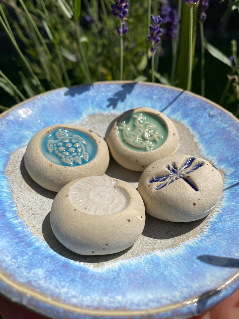 May include: A blue ceramic dish holds four small, round, off-white ceramic stones. Each stone has a different embossed design: a turtle, a seahorse, a dragonfly, and a spiral. The dish has a blue and gray glaze.
