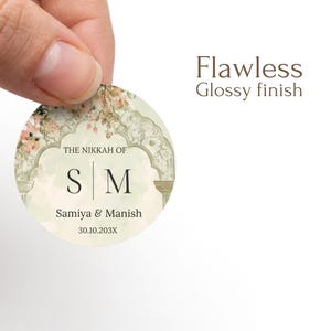 May include: A circular sticker with a glossy finish featuring an arched architectural design, floral accents, and the text THE NIKKAH OF, S M, Samiya & Manish, and 30.10.203X. The sticker has a light cream background with soft green and pink tones.