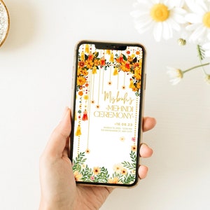 May include: A smartphone displays a digital invitation for a Mehndi ceremony. The design features floral arrangements in yellow and orange, with hanging tassels and the event details in gold script. The phone is held in a hand.