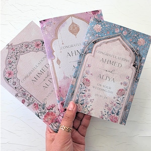 May include: Three rectangular wedding cards featuring an arched frame design with floral patterns in shades of pink, blue, and gold. The text reads: CONGRATULATIONS AHMED and ALIYA ON YOUR WEDDING DAY 25 | 10 | 2X. The cards have a textured paper finish.