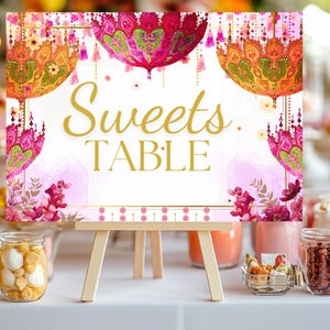 May include: A wooden easel displays a sign that reads "Sweets TABLE" in gold script. The sign features ornate, colorful designs with pink, orange, and green hues. The table is set with various jars and containers filled with treats.