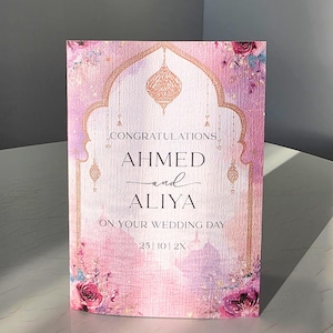 May include: A rectangular wedding card with a pink and purple watercolor background featuring a gold arched frame, hanging lanterns, and floral accents. The text reads: CONGRATULATIONS AHMED and ALIYA ON YOUR WEDDING DAY 25 | 10 | 2X in black lettering.