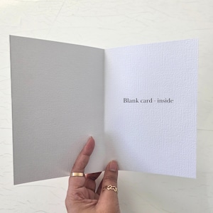 May include: A white greeting card with a textured surface, shown open to reveal a blank interior. The text "Blank card - inside" is printed in black on the right side of the card. The card is held open, displaying its clean, simple design.