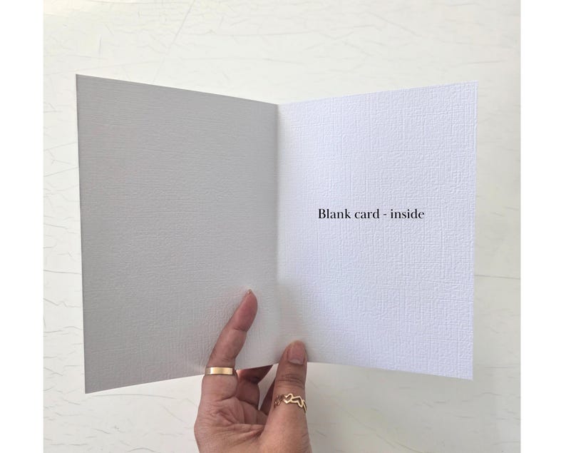 May include: A white greeting card with a textured surface, shown open to reveal a blank interior. The text "Blank card - inside" is printed in black on the right side of the card. The card is held open, displaying its clean, simple design.