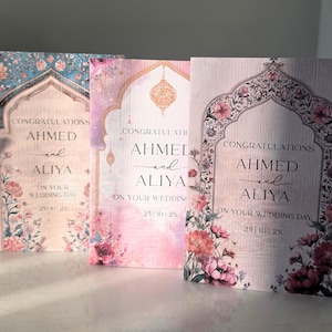 May include: Three rectangular wedding cards featuring an arched frame design with floral accents. Each card displays the text CONGRATULATIONS AHMED and ALIYA ON YOUR WEDDING DAY 25 | 10 | 2X. The cards feature blue, pink, and light purple color palettes.