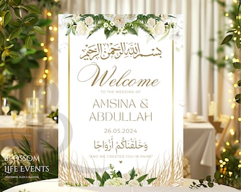 May include: A white wedding welcome sign with gold accents and floral decorations. The sign reads "Bismillah ar-Rahman ar-Rahim" in Arabic script, followed by "Welcome" in a cursive font. The names "Amsina & Abdullah" are listed below, along with the date "26.05.2024". The sign also includes the Arabic phrase "Wa khalaqnakum azwajan" and the English translation "And we created you in pairs".