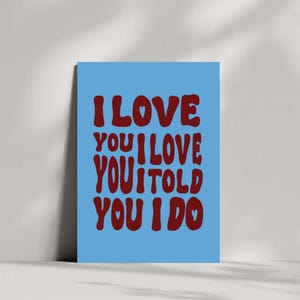 May include: A blue poster with the words "I LOVE YOU I LOVE YOU I TOLD YOU I DO" in a bold, red font. The word "LOVE" has a heart symbol replacing the letter "O". The poster is leaning against a white wall.