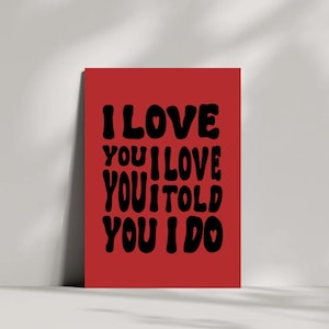 May include: A red rectangular art print with the words "I LOVE YOU I LOVE YOU I TOLD YOU I DO" in black, bold, rounded font. The print has a white border and is leaning against a white wall.