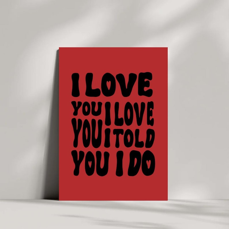 May include: A red rectangular art print with the words "I LOVE YOU I LOVE YOU I TOLD YOU I DO" in black, bold, rounded font. The print has a white border and is leaning against a white wall.