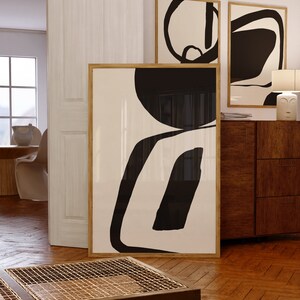 K&ouml;nnte beinhalten: Abstrakte Kunstdrucke in Holzrahmen. Das Kunstwerk zeigt kr&auml;ftige schwarze Formen auf cremefarbenem Hintergrund. Der gr&ouml;&szlig;te Druck lehnt an einer Wand, w&auml;hrend zwei kleinere Drucke &uuml;ber einem Holzschrank h&auml;ngen. Der Raum hat Parkettb&ouml;den.