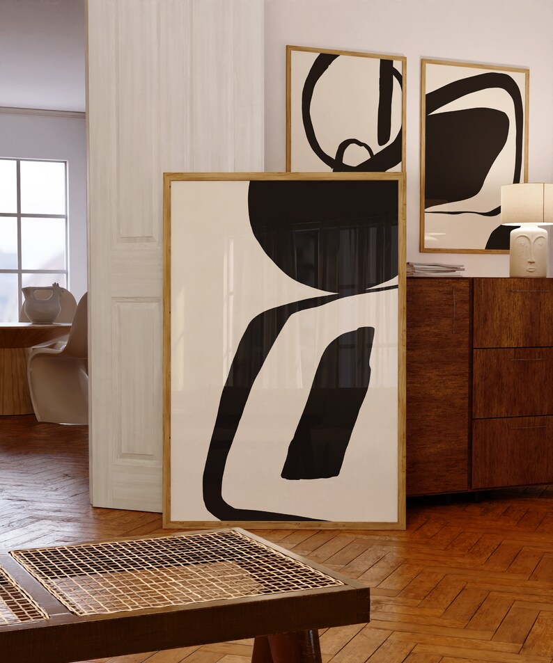 K&ouml;nnte beinhalten: Abstrakte Kunstdrucke in Holzrahmen. Das Kunstwerk zeigt kr&auml;ftige schwarze Formen auf cremefarbenem Hintergrund. Der gr&ouml;&szlig;te Druck lehnt an einer Wand, w&auml;hrend zwei kleinere Drucke &uuml;ber einem Holzschrank h&auml;ngen. Der Raum hat Parkettb&ouml;den.