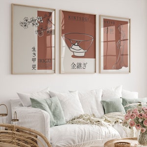 Op de afbeelding: Drie ingelijste prints met Japanse woorden en illustraties. De eerste print toont een tak met kersenbloesems met de woorden "IKIGAI" en "生き甲斐". De tweede print toont een gebroken theekopje met gouden accenten en de woorden "KINTSUGI" en "金継ぎ". De derde print toont een vrouw in een kimono met de woorden "UKIYO" en "浮世".