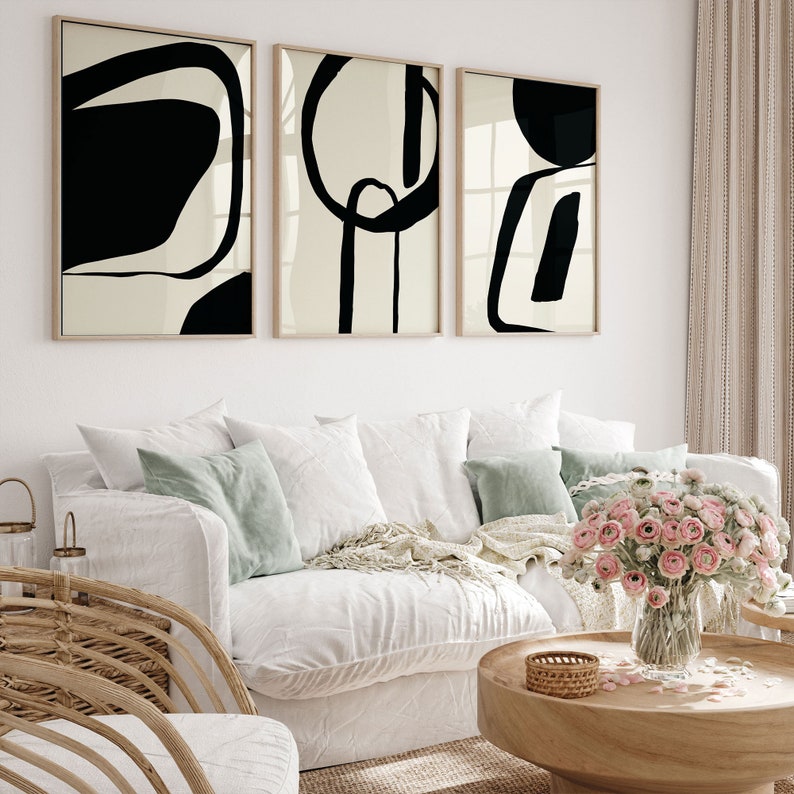 K&ouml;nnte beinhalten: Drei gerahmte abstrakte Kunstdrucke mit schwarzen Formen auf cremefarbenem Hintergrund h&auml;ngen &uuml;ber einem wei&szlig;en Sofa mit gr&uuml;nen und wei&szlig;en Kissen. Ein Holztisch mit einem geflochtenen Korb und einer Vase mit rosa Blumen vervollst&auml;ndigt das Wohnzimmer.