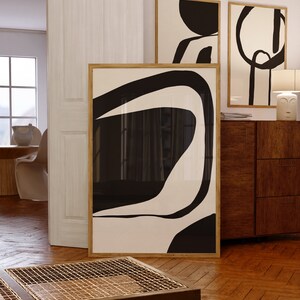 K&ouml;nnte beinhalten: Abstrakte Kunstdrucke in Holzrahmen. Das Kunstwerk zeigt kr&auml;ftige schwarze Formen auf cremefarbenem Hintergrund. Die Drucke werden in einem Raum mit Parkettboden und einem Holzschrank ausgestellt.