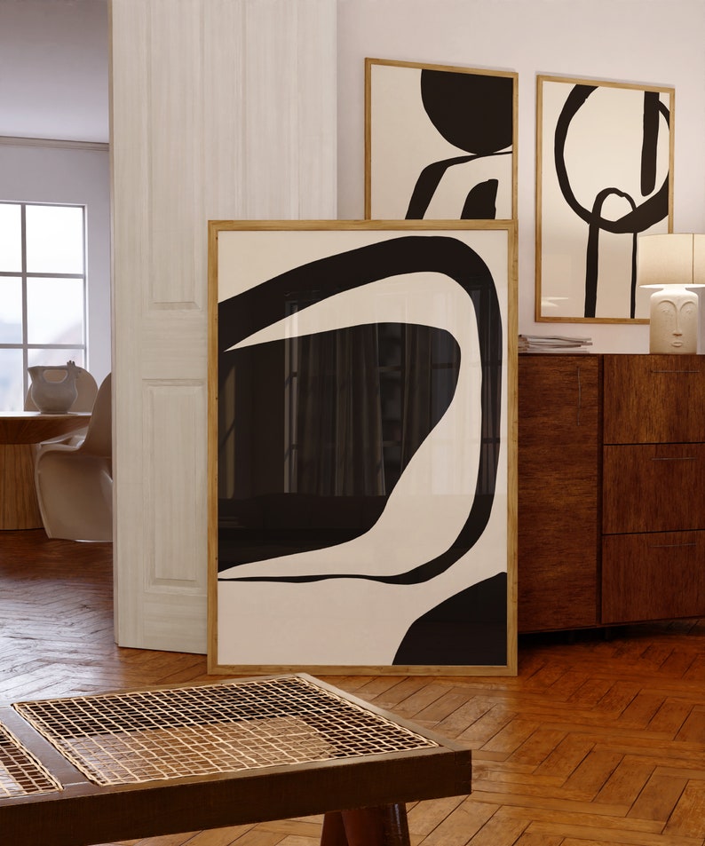 K&ouml;nnte beinhalten: Abstrakte Kunstdrucke in Holzrahmen. Das Kunstwerk zeigt kr&auml;ftige schwarze Formen auf cremefarbenem Hintergrund. Die Drucke werden in einem Raum mit Parkettboden und einem Holzschrank ausgestellt.