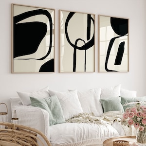 K&ouml;nnte beinhalten: Drei gerahmte abstrakte Kunstdrucke. Jeder Druck zeigt schwarze Formen auf cremefarbenem Hintergrund mit einem hellen Holzrahmen. Das Kunstwerk wird &uuml;ber einem wei&szlig;en Sofa mit Kissen und einem Holztisch ausgestellt.