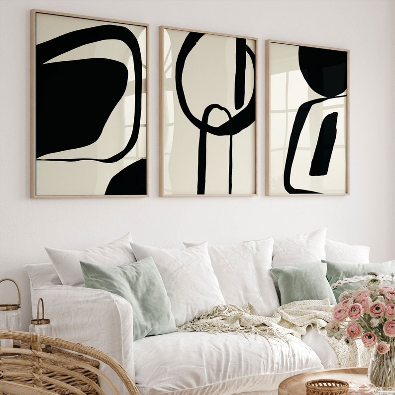 K&ouml;nnte beinhalten: Drei gerahmte abstrakte Kunstdrucke. Jeder Druck zeigt schwarze Formen auf cremefarbenem Hintergrund mit einem hellen Holzrahmen. Das Kunstwerk wird &uuml;ber einem wei&szlig;en Sofa mit Kissen und einem Holztisch ausgestellt.