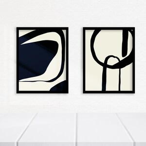 K&ouml;nnte beinhalten: Zwei gerahmte abstrakte Kunstdrucke. Einer zeigt ein marineblaues und cremefarbenes Design, der andere ein schwarz-cremefarbenes Design. Beide sind in schwarzen Rahmen und werden auf einer wei&szlig;en Oberfl&auml;che pr&auml;sentiert.