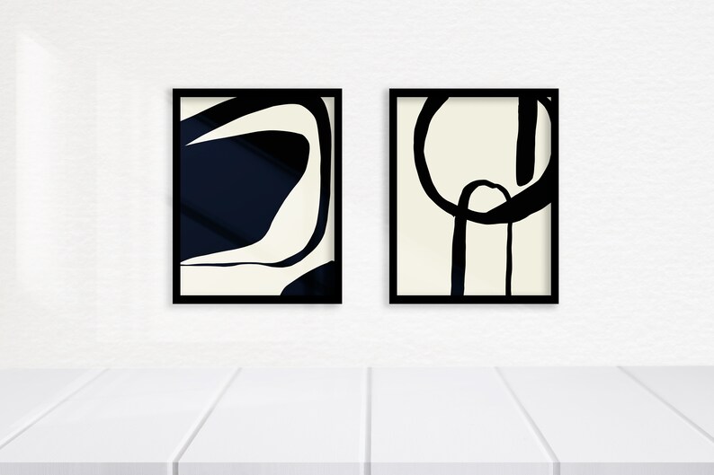 K&ouml;nnte beinhalten: Zwei gerahmte abstrakte Kunstdrucke. Einer zeigt ein marineblaues und cremefarbenes Design, der andere ein schwarz-cremefarbenes Design. Beide sind in schwarzen Rahmen und werden auf einer wei&szlig;en Oberfl&auml;che pr&auml;sentiert.
