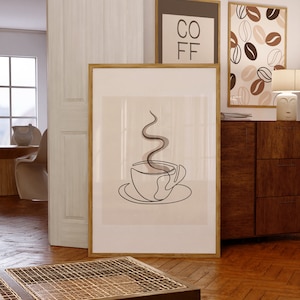 以下が含まれることがあります： コーヒーカップとそこから立ち上る蒸気を描いた線画の額装されたプリント。プリントは薄いベージュの背景にされており、木製の額縁が施されています。プリントは、他の2つの額装されたプリントの隣に白い壁にかけられています。