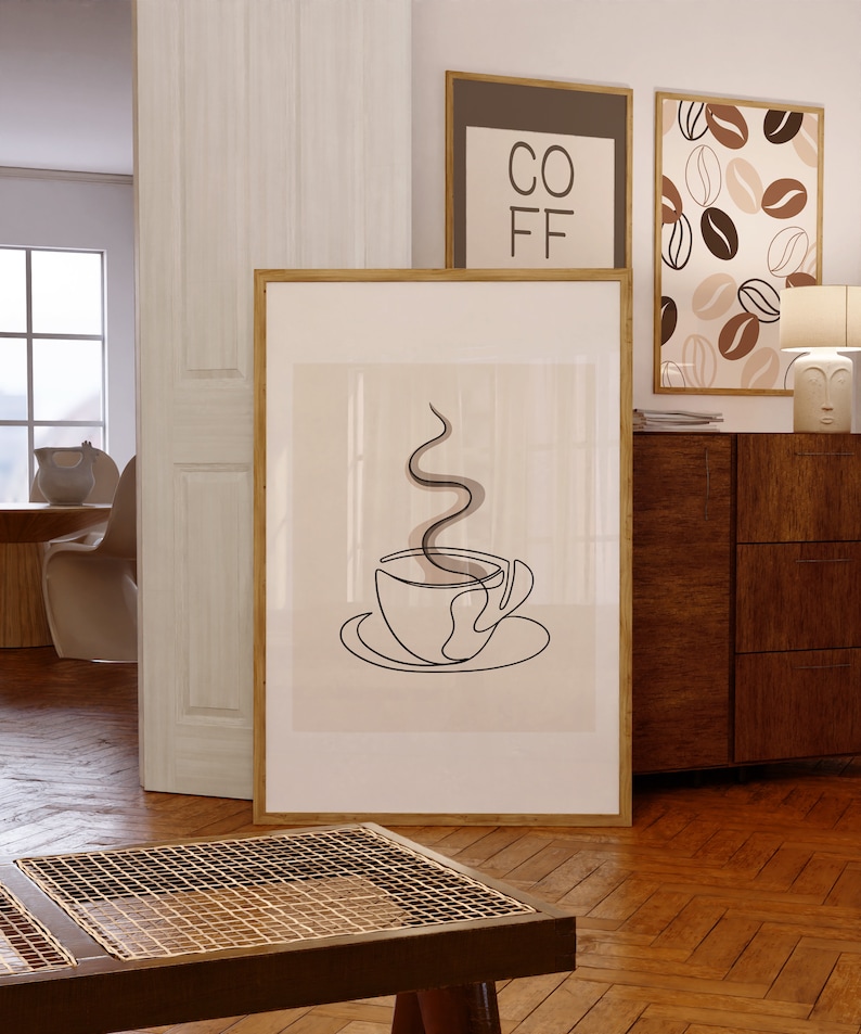 以下が含まれることがあります： コーヒーカップとそこから立ち上る蒸気を描いた線画の額装されたプリント。プリントは薄いベージュの背景にされており、木製の額縁が施されています。プリントは、他の2つの額装されたプリントの隣に白い壁にかけられています。