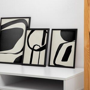 K&ouml;nnte beinhalten: Drei gerahmte abstrakte Kunstdrucke mit schwarzen und cremefarbenen Designs. Die Kunstwerke zeigen kr&auml;ftige, grafische Formen und Linien. Die Rahmen sind schwarz und lehnen an einem wei&szlig;en Schreibtisch. Eine Vase mit einer getrockneten Pflanze und ein Holzregal mit B&uuml;chern sind ebenfalls zu sehen.