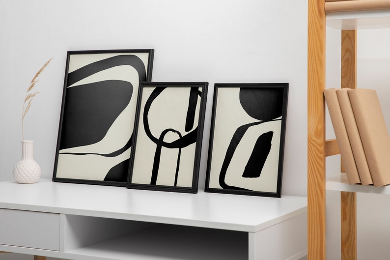K&ouml;nnte beinhalten: Drei gerahmte abstrakte Kunstdrucke mit schwarzen und cremefarbenen Designs. Die Kunstwerke zeigen kr&auml;ftige, grafische Formen und Linien. Die Rahmen sind schwarz und lehnen an einem wei&szlig;en Schreibtisch. Eine Vase mit einer getrockneten Pflanze und ein Holzregal mit B&uuml;chern sind ebenfalls zu sehen.