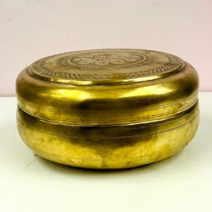 Könnte beinhalten: Eine runde, goldfarbene Metallbox mit Deckel. Der Deckel hat ein geprägtes Blumenmuster. Die Box steht auf einer weißen Oberfläche mit dekorativem Holzbesatz.