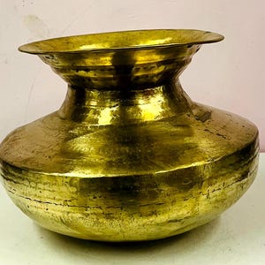 May include: A golden-colored, antique-style brass pot with a wide, rounded body and a flared rim. The pot has a textured surface and a slightly uneven shape, suggesting it is handmade. The pot is displayed on a white surface.